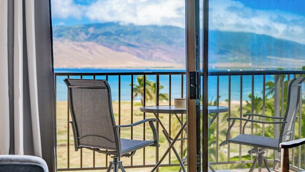A balcony with two chairs and a small table, overlooking a beach and mountains.