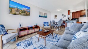A modern living room with a sofa, a coffee table, and a TV mounted on the wall. There is a dining area with a table and chairs, and a kitchen area with wooden cabinets and a countertop.