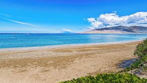A beach with clear blue water, a sandy shore, and distant mountains under a blue sky.