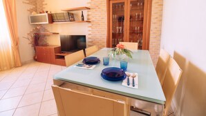 Una zona de menjador amb una taula de vidre preparada per a dues persones, una paret de maó, un armari de fusta amb portes de vidre i una televisió sobre una estantania.