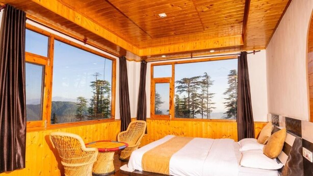 A wooden cabin room with a bed, two wicker chairs, a small table, and large windows offering a mountain view. - Ahistay Homes, Fagu (Fagu)