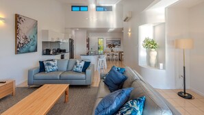 A modern living room with a grey sofa, patterned rug, and wooden coffee table. A kitchen and dining area are visible in the background. - Anchorage 3, Anchorage 3 on Hamilton Island (Hamilton Island)
