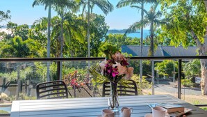 A table with a vase of flowers, two cups, and a book on a balcony overlooking a tropical landscape with palm trees and a view of the sea. - Anchorage 3, Anchorage 3 on Hamilton Island (Hamilton Island)