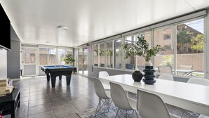 Un vaste espace intérieur comprenant une table de billard, une table à manger avec des chaises, et de grandes fenêtres donnant sur l’extérieur.