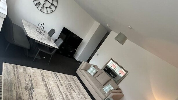 A modern living room with a gray sofa, a dining area with a table and chairs, and a large wall clock. - The M Rooms (Shotts)