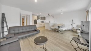 Ein modernes Wohnzimmer mit einem grauen Sofa, einem runden Couchtisch und einem Essbereich mit weißen Stühlen und einem kleinen Tisch.