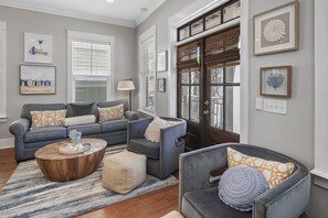 Un salon avec un canapé gris, une table basse et un fauteuil. Des tableaux encadrés ornent le mur, et une porte avec une fenêtre est également présente.