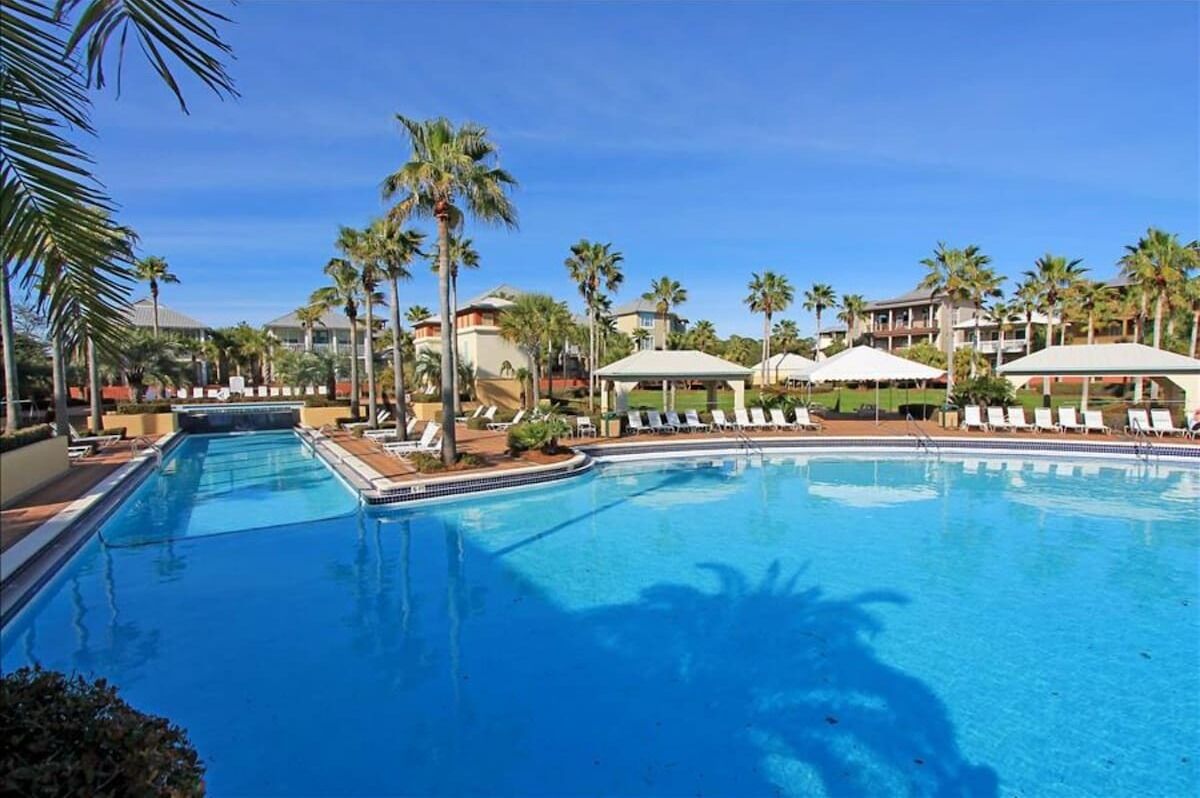 Ein Swimmingpool mit Liegestühlen und Sonnenschirmen, umgeben von Palmen und Hotelgebäuden.