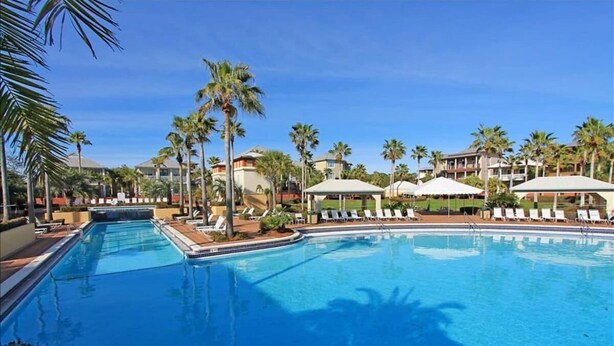 Una piscina con sillas de descanso y sombrillas, rodeada de palmeras y edificios de hotel.