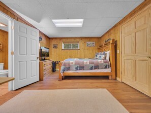 Un dormitorio acogedor con piso de madera, una cama con colcha, un aparador, un televisor y una ventana.