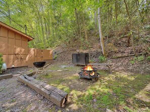 Un espacio exterior rústico con una cabaña de madera, un pozo de fuego y una parrilla de metal.