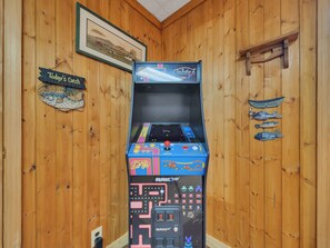 Una máquina de arcade retro en una habitación con paneles de madera, un cuadro de paisaje enmarco y un letrero que dice 'Hoy en el menú'.