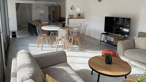 Uma sala de estar moderna com sofá, mesa de centro e televisão. Há uma área de jantar com cadeiras e mesa. O cômodo possui teto de madeira e paredes brancas.
