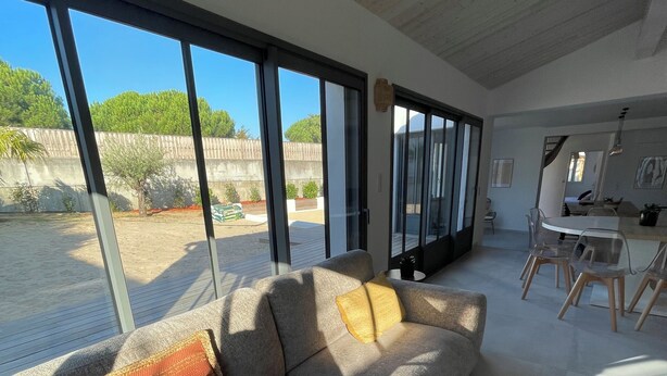 Uma sala de estar moderna com um sofá cinzento, uma mesa de centro de madeira e grandes janelas com vista para um quintal.