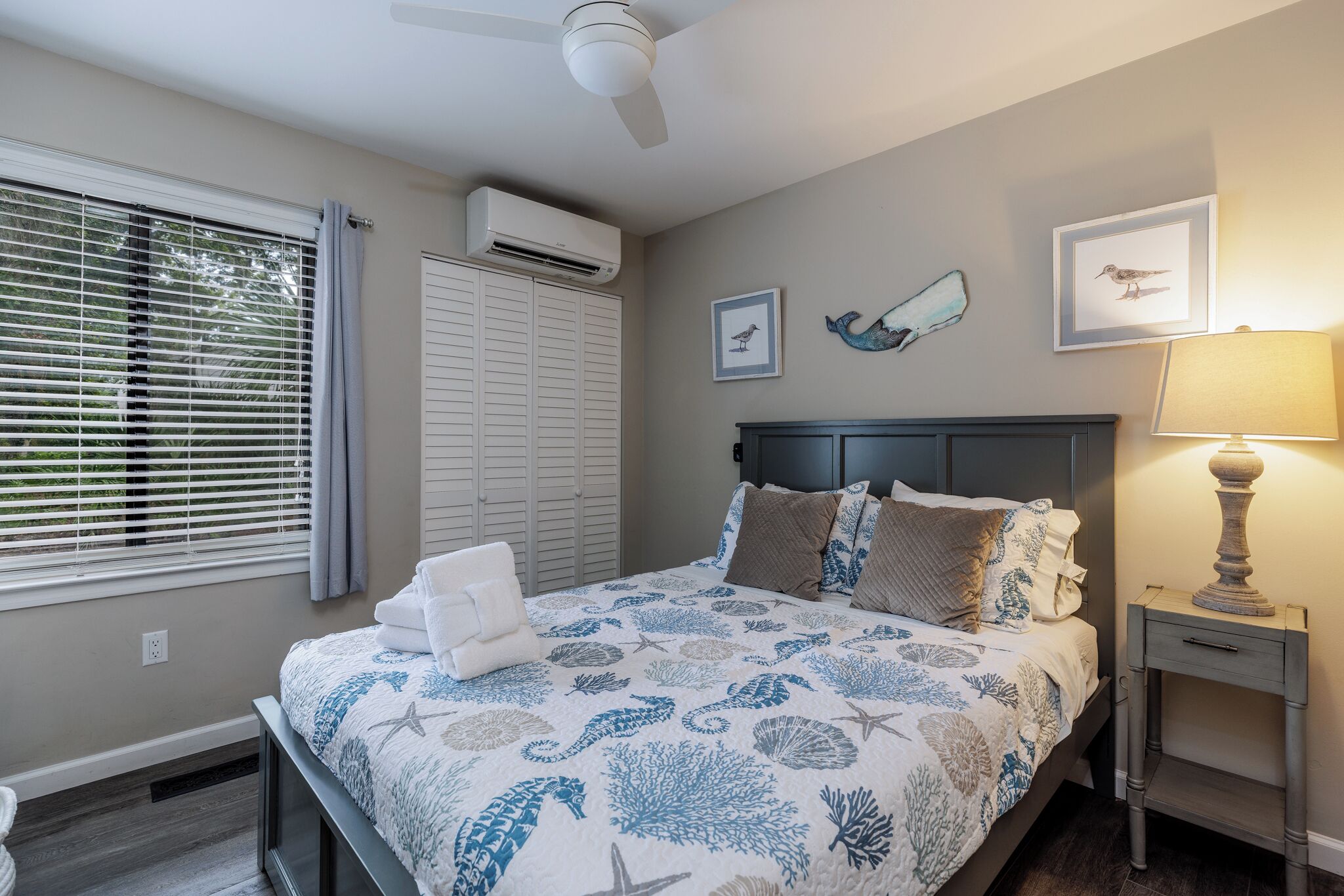 Un dormitorio con cama, mesita de noche, lámpara, ventilador de techo, unidad de aire acondicionado y ventana con persianas.