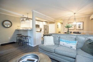 Cocina moderna con armarios blancos, microondas y refrigerador. Hay una zona de comedor con un sofá gris y una mesa de centro.