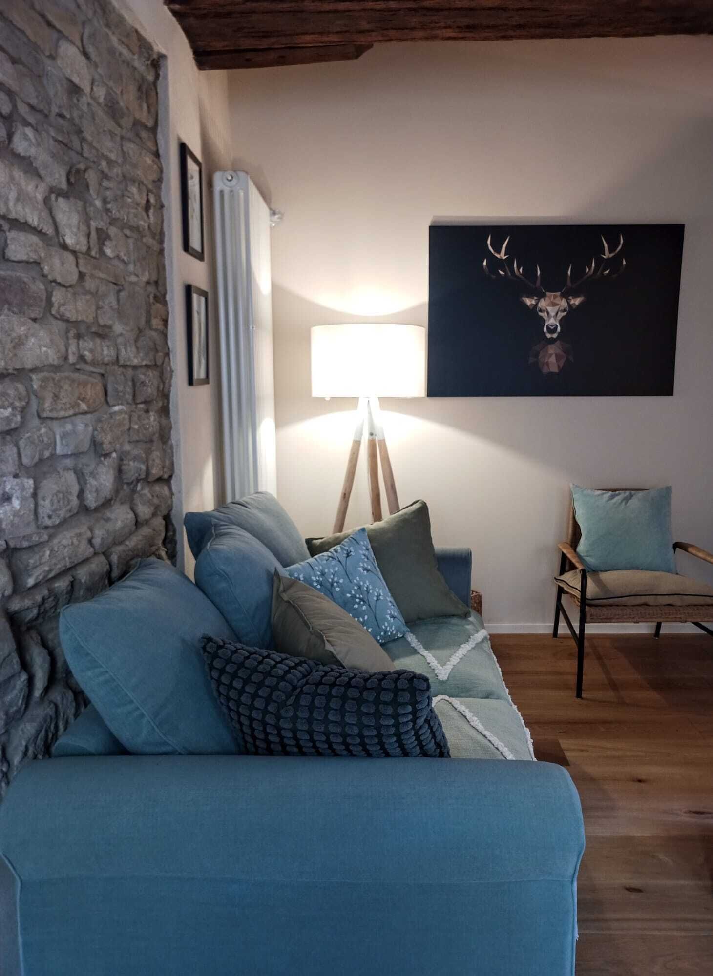 A cozy living room with a stone wall, a blue sofa with patterned cushions, a wooden floor, and a deer-themed artwork on the wall.
