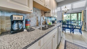 A modern kitchen with granite countertops, a coffee maker, and a sink. There is a dining area with blue chairs and a chandelier. - New to Seascape Vacations in a fantastic location! Small dogs allowed! (Miramar Beach)