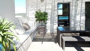 A sunlit outdoor seating area with a potted plant, a cushioned bench, and a glass table.