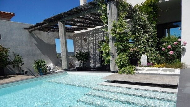 A swimming pool with clear water, surrounded by a pergola, plants, and a white building.