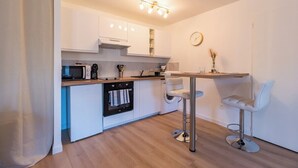 A modern kitchen with white cabinets, a built-in oven, a microwave, and a coffee maker. There is a small dining area with a table and two white chairs. - Studio Cosy - plein coeur de Lannion (Lannion)