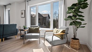 Ein modernes Wohnzimmer mit einer Couch, zwei Sesseln, einem Couchtisch und einem großen Fenster mit Blick auf Häuser.