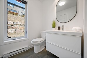 A modern bathroom with a toilet, a white vanity, a mirror, and a window with a view of a stone wall and a building. - Maison Oasis by RVMT (Mont-Blanc)