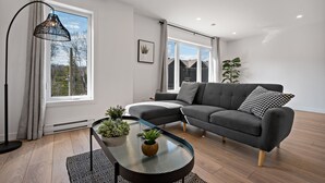 Ein modernes Wohnzimmer mit einem grauen Ecksofa, einem Glastisch mit Pflanzen, einer schwarzen Stehlampe und großen Fenstern mit Vorhängen.