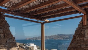 A stone-walled room with a view of the sea, a round table, wicker chairs, and a wooden ceiling structure. - Ocean whisper villa with amazing view  (mykonos)