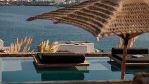 A poolside area with lounge chairs and a woven umbrella, overlooking a body of water and a distant cityscape. - Ocean whisper villa with amazing view  (mykonos)