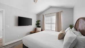 Une chambre avec un grand lit, un ventilateur de plafond, un téléviseur fixé au mur, une fenêtre avec des stores et une plante en pot.