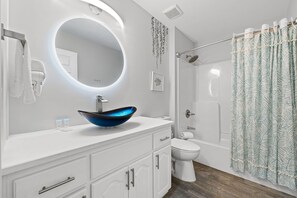 A modern bathroom with a round mirror, a blue vessel sink, a toilet, and a shower with a leaf-patterned curtain.