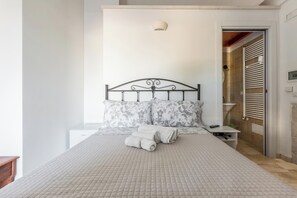 Una cama bien hecha con un cubrecolchón acolchado, dos almohadas estampadas con motivos florales y un juego de toallas dobladas sobre la cama.