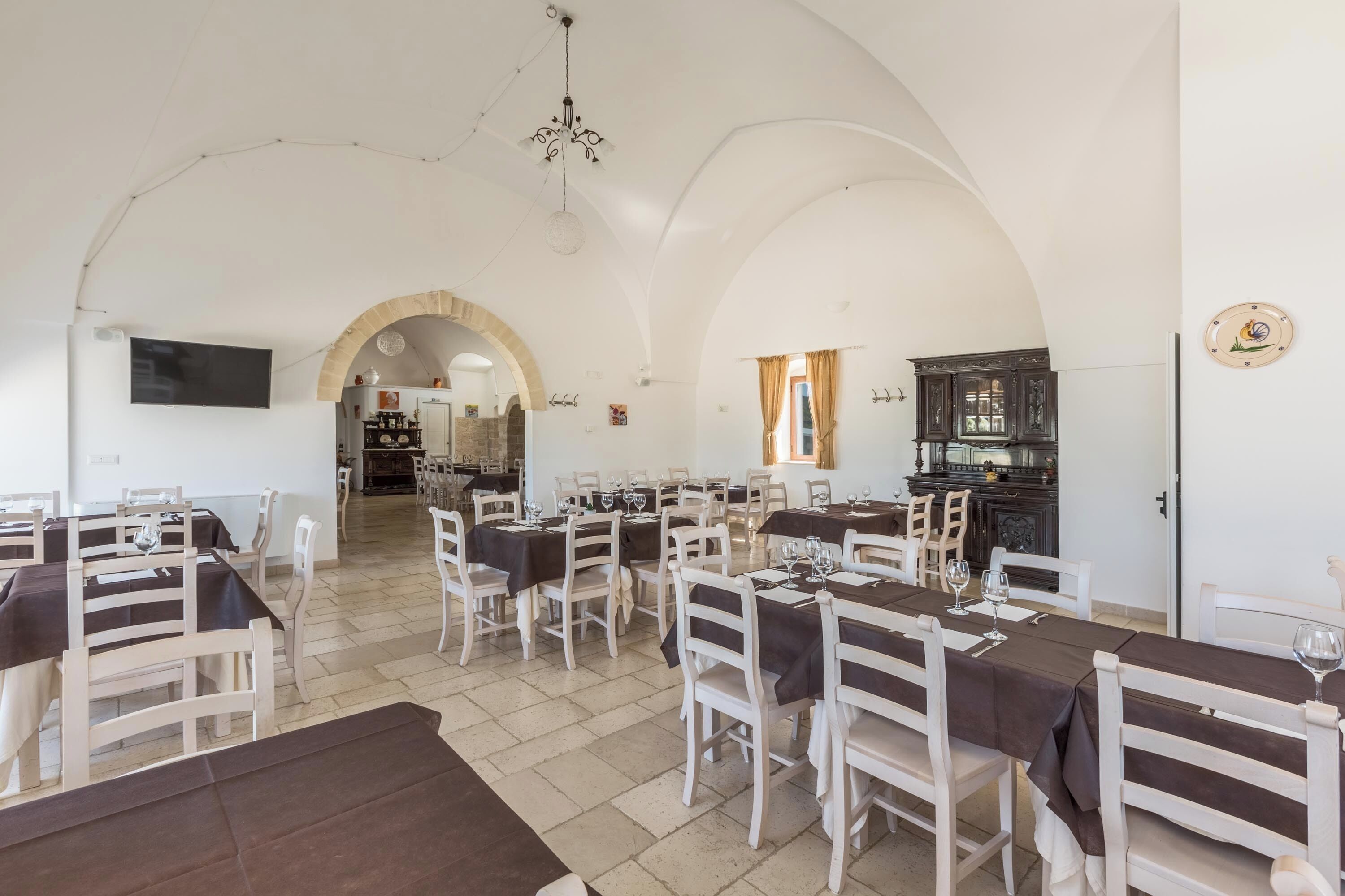 Un comedor espacioso con techos altos en arco, paredes blancas y un arco central que da a otra habitación. Las mesas están dispuestas con sillas blancas y manteles oscuros, y un candelabro cuelga del techo.