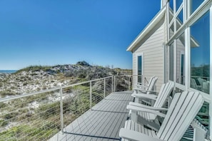 A beach house with a balcony, white chairs, and a view of the beach and dunes.