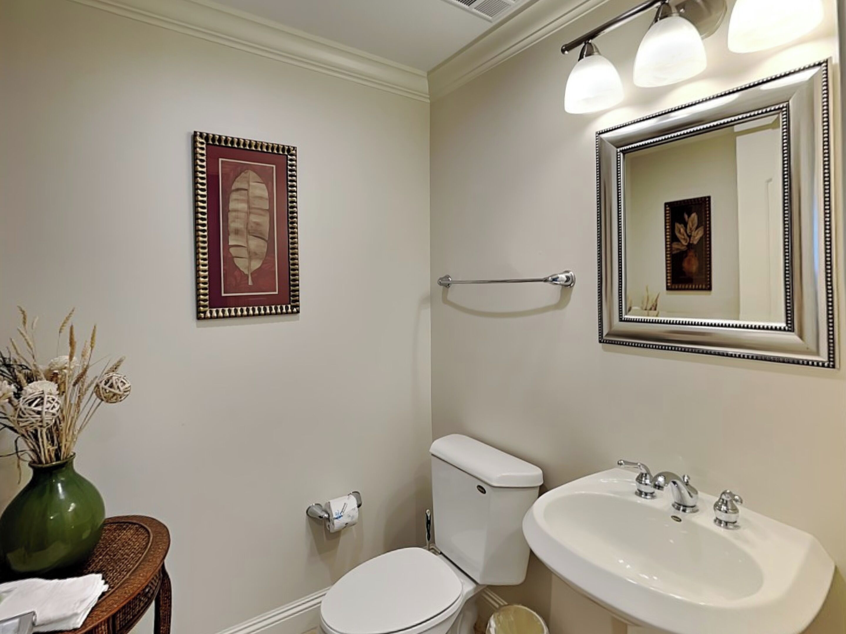 Une salle de bain avec des toilettes, un lavabo et un miroir. Un tableau encadré est accroché au mur et un vase contenant des plantes séchées est posé sur une table.