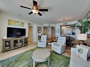 A spacious living room with a flat-screen TV, white armchairs, a round coffee table, and a ceiling fan. - Indigo East #1203 | Gulf Front, Breathtaking Views + Resort Amenities! (Perdido Key)