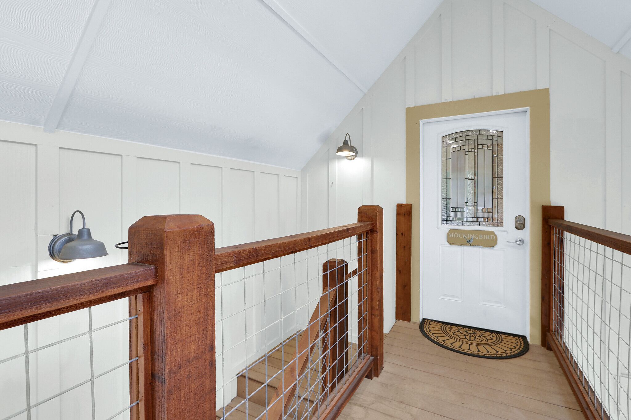 Un escalier en bois avec une rampe en métal menant à une porte blanche dotée d’un panneau de verre et surmontée d’un paillasson.