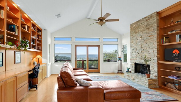 A spacious living room with a leather sofa, wooden bookshelves, a fireplace, and large windows offering a view of the outdoors. - Casa Robles- Mockingbird I Couples, stargazing (Wimberley)