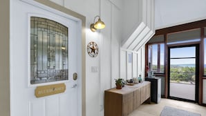 A hallway with a white door labeled "BLUEBONNET," a wooden bench, a wall-mounted light fixture, and a large window with a view. - Casa Robles- Bluebonnet I Firepit, couples (Wimberley)