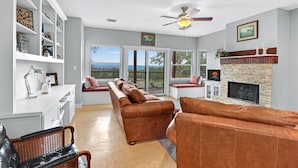 A spacious living room with a fireplace, leather sofas, a bookshelf, and large windows offering a view of the outdoors. - Casa Robles- Bluebonnet I Firepit, couples (Wimberley)