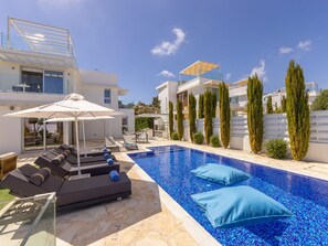 Ein moderner Poolbereich im Freien mit Liegestühlen und Sonnenschirmen, umgeben von hohen Zypressen und einem strahlend blauen Himmel.