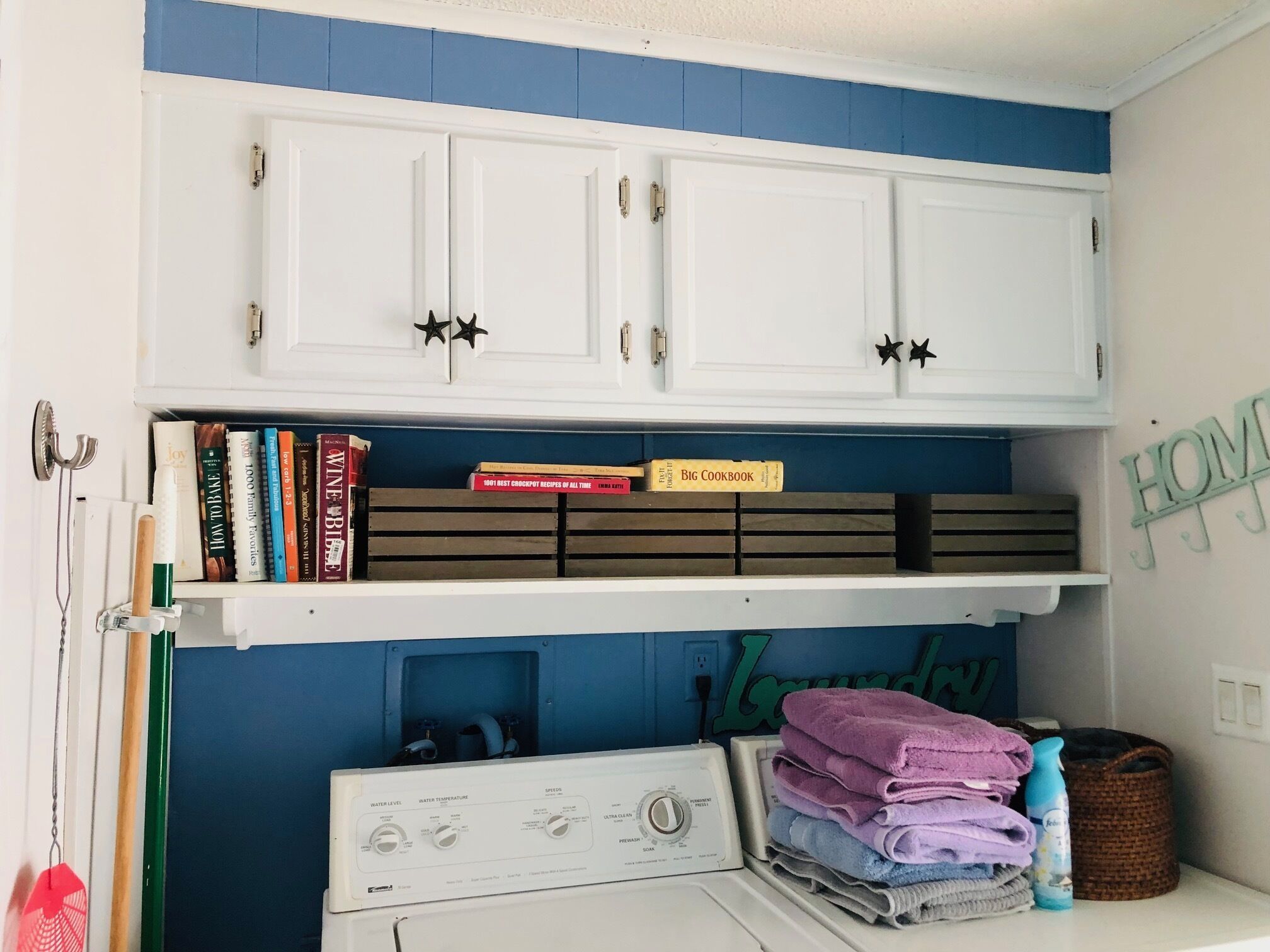 A laundry area with a washing machine, stacked towels, and a shelf with books and a 'Big Cookbook'.