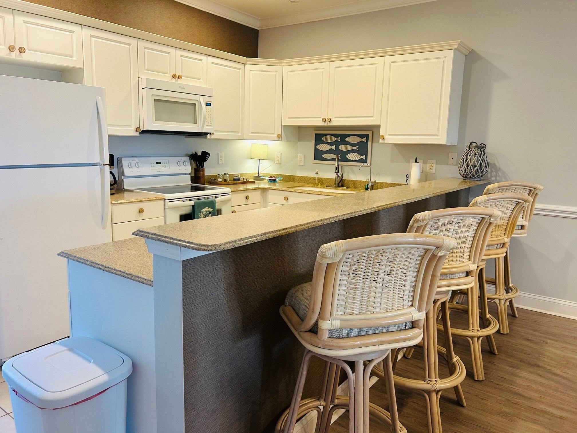 A kitchen with white cabinets, a microwave, a stove, a sink, and a bar area with wicker chairs.