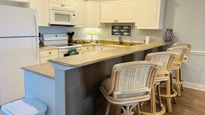A kitchen with white cabinets, a microwave, a stove, a sink, and a bar area with wicker chairs.