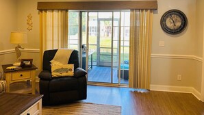 A living room with a black armchair, a lamp, a clock, and a 'Beach' sign above a sliding glass door leading to a patio.