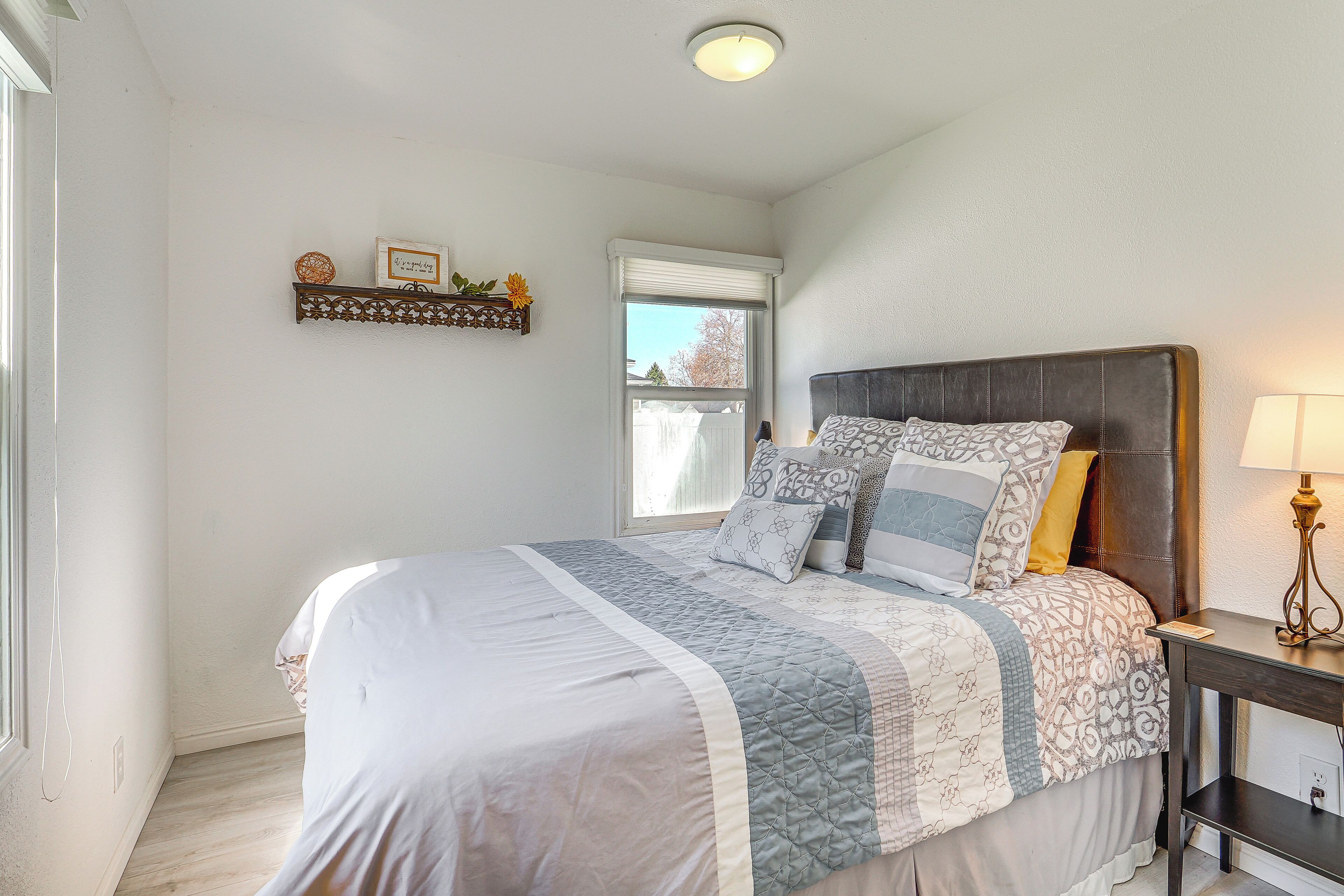 A bedroom with a large bed, a bedside table with a lamp, a window with blinds, and a shelf with decorative items.