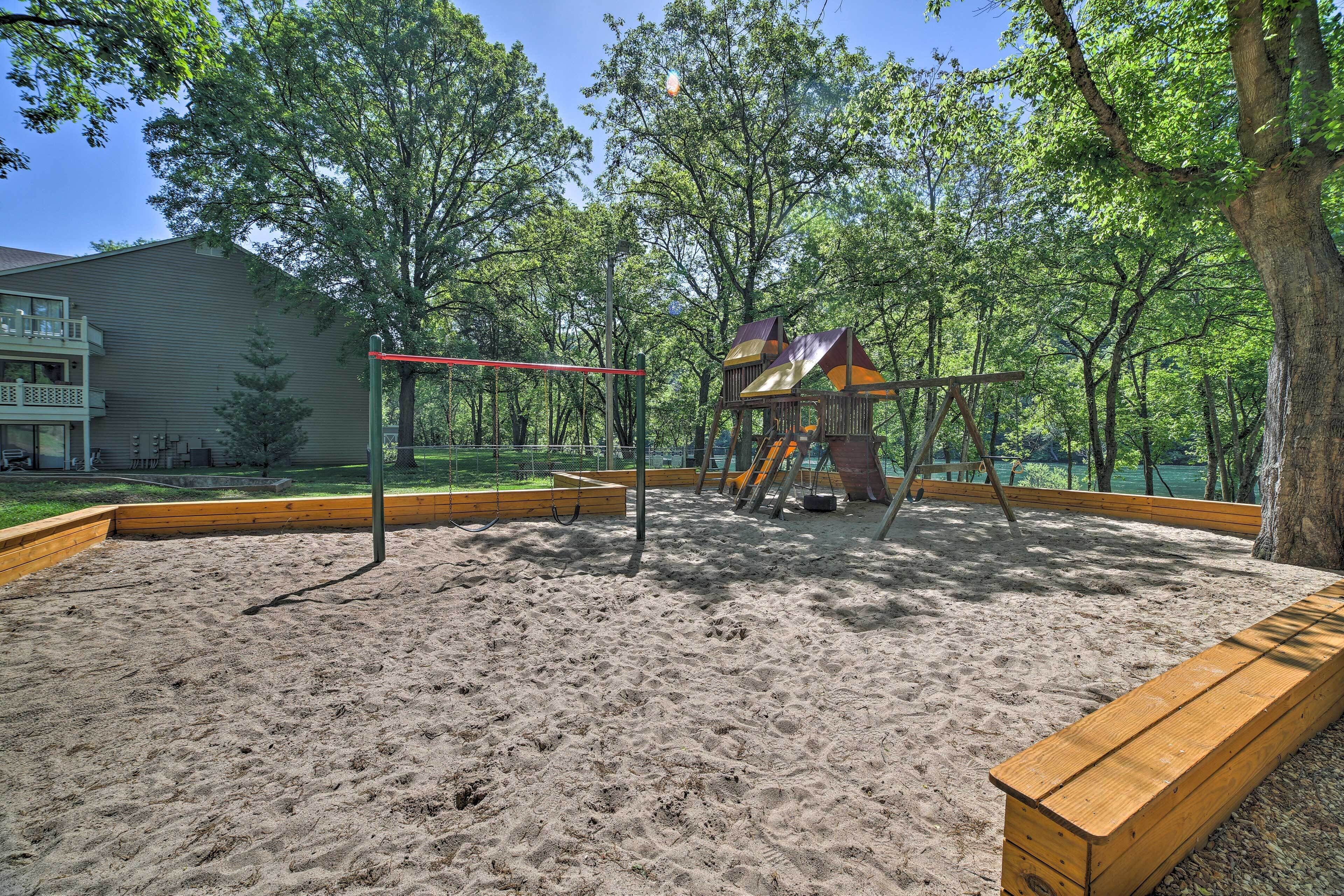 A playground with a swing set and a climbing structure surrounded by sand and trees.