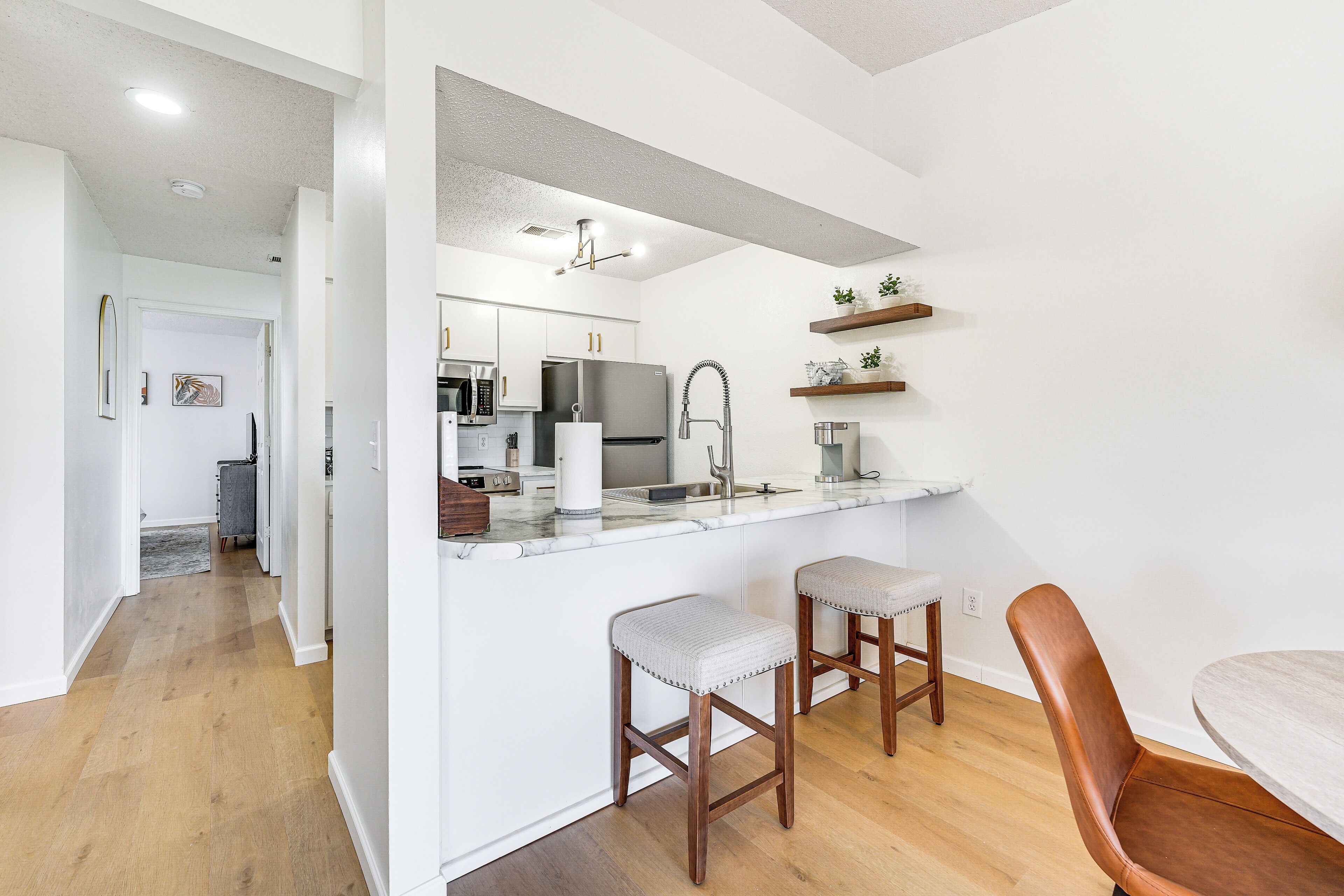 Una cocina moderna con armarios blancos, electrodomésticos de acero inoxidable y encimera de mármol. Hay dos taburetes de madera y una silla marrón.