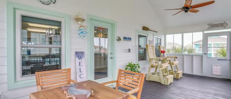 A covered porch with a wooden table and chairs, a ceiling fan, and a view of the house in the background.